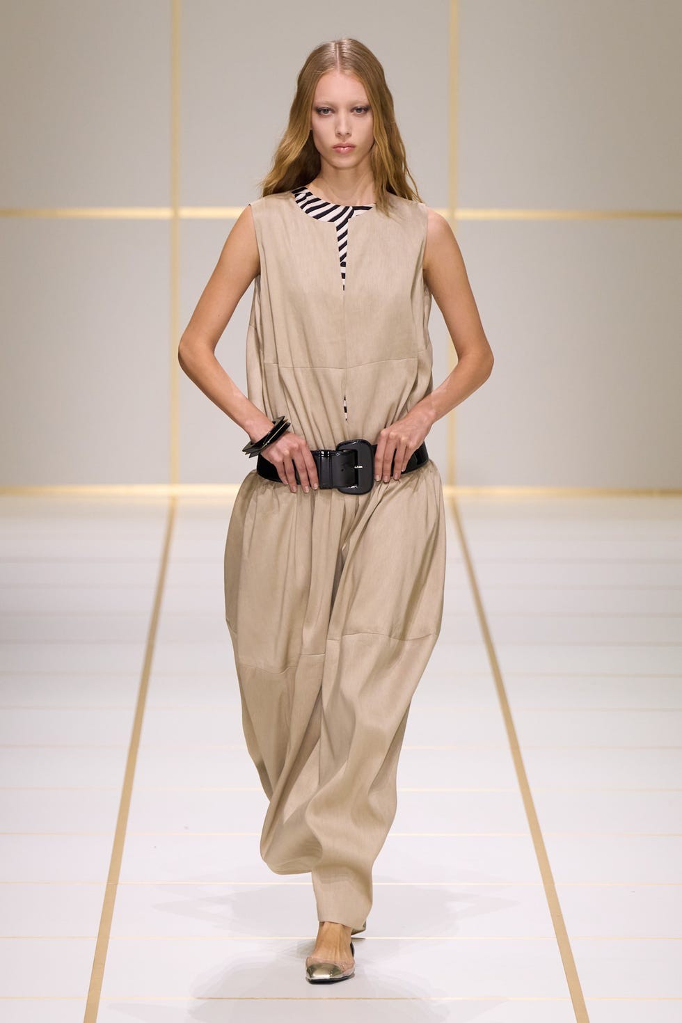 A model walks the runway in a sleeveless, beige jumpsuit with a black belt and striped inner top at a fashion show.