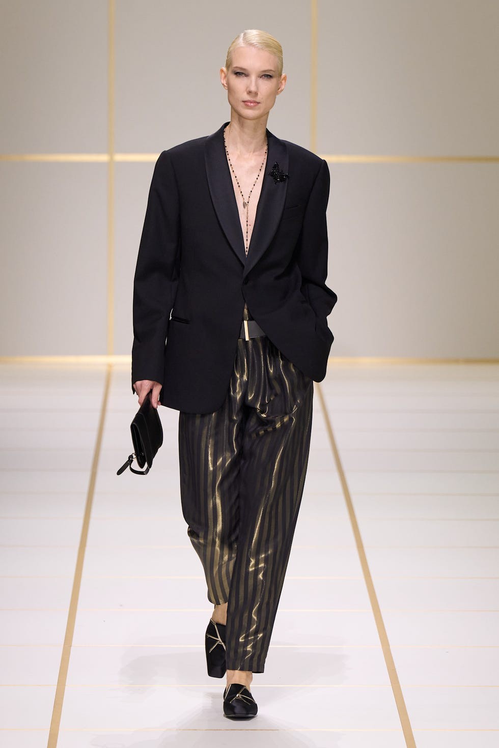 Model wearing a black blazer and striped pants on a fashion runway with a minimalist background.