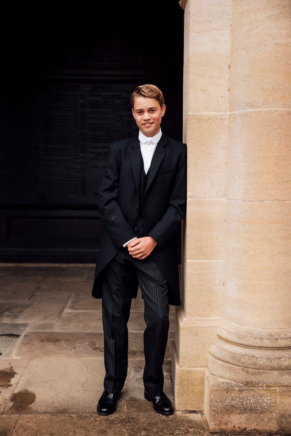 Young man in formal attire leaning against a stone column, smiling confidently.