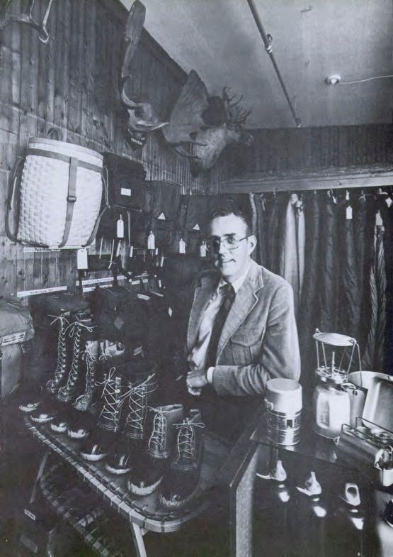 A man in glasses stands in a store displaying various outdoor gear, including boots and camping equipment.