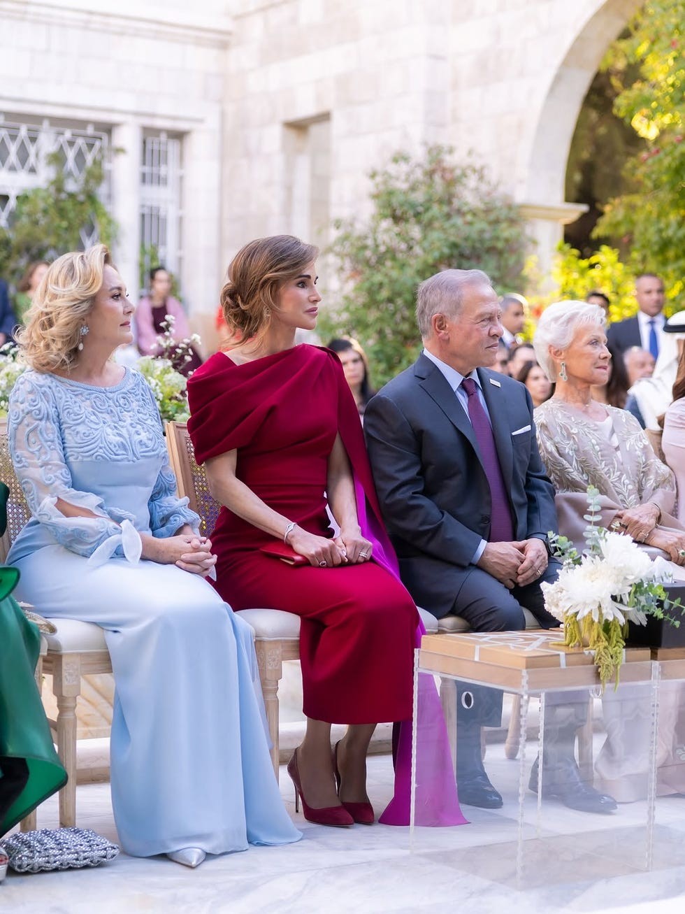 Four elegantly dressed individuals seated together at a formal event, surrounded by greenery and a gathering audience.