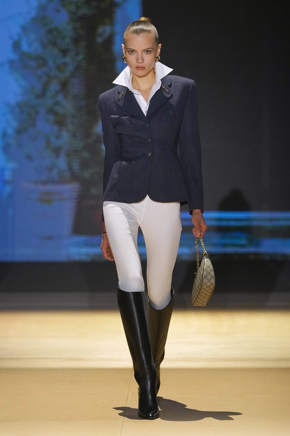 Model walking the runway in a stylish navy blazer, white pants, and black knee-high boots, holding a handbag.