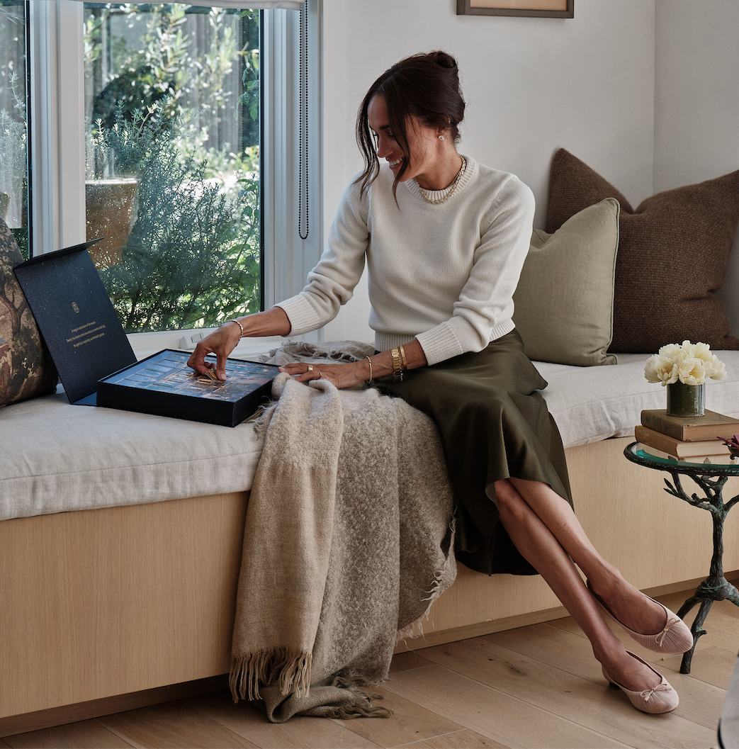 A woman sitting on a bench, examining a box while wrapped in a blanket, with plants and furnishings in a bright room.