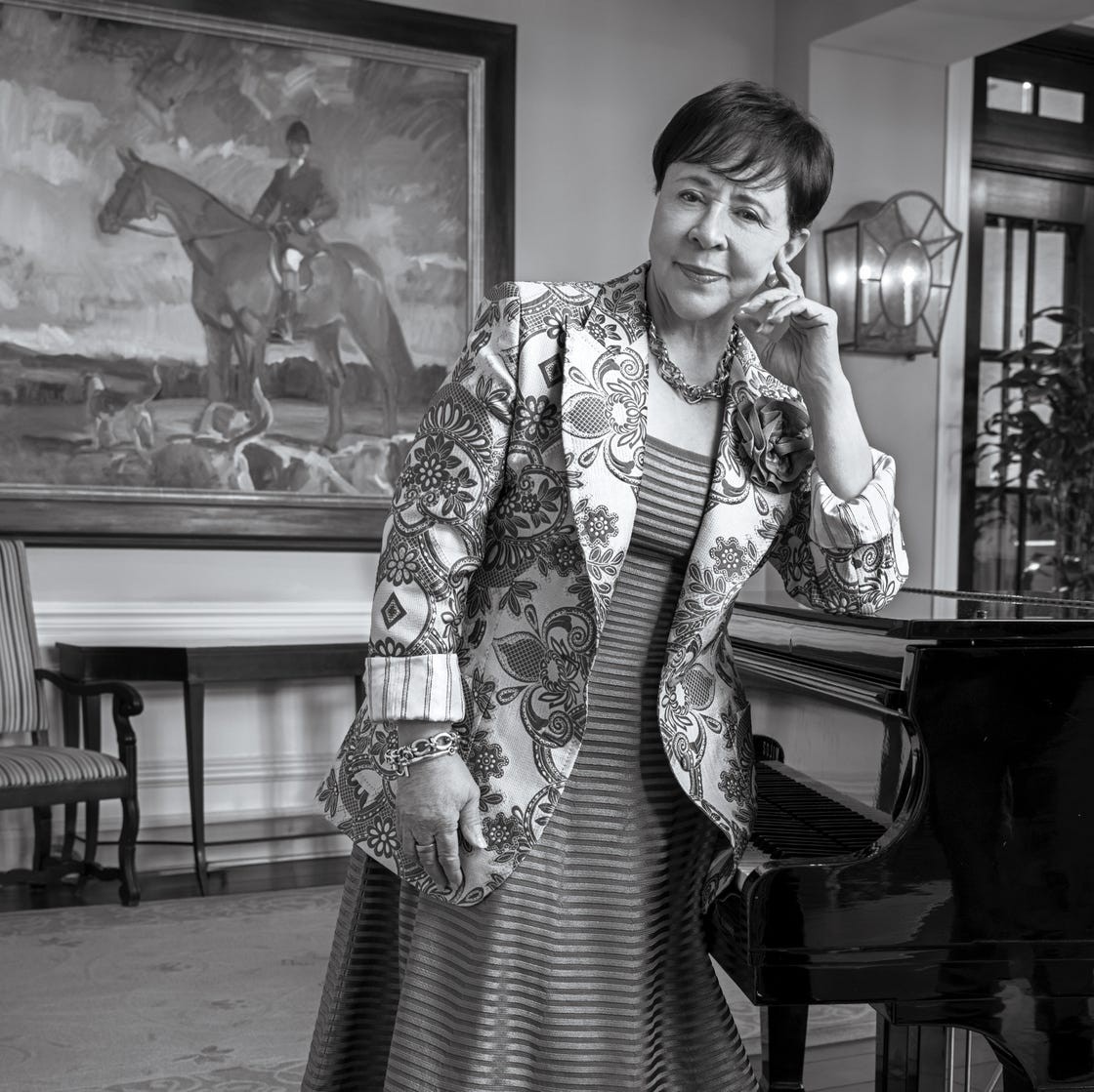 A woman in a decorative jacket and dress poses beside a piano in an elegantly decorated room with a horse painting.