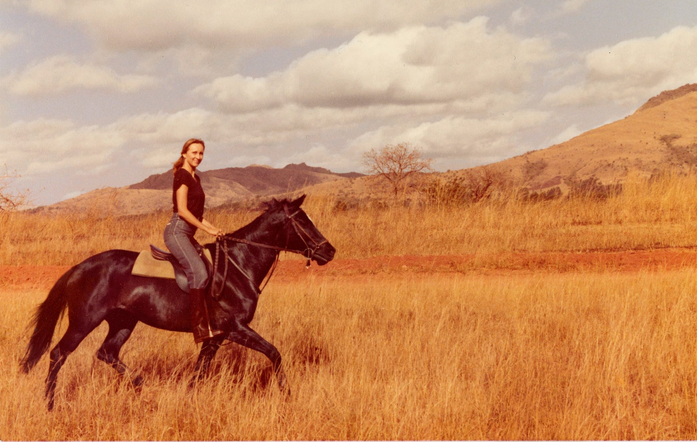 Klara Glowczewska A rider on a black horse gallops across a grassy landscape with mountains in the background under a cloudy sky.