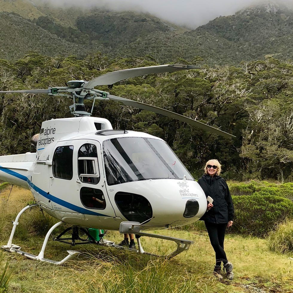 Klara Glowczewska A person stands beside a helicopter in a mountainous area with trees and grass.
