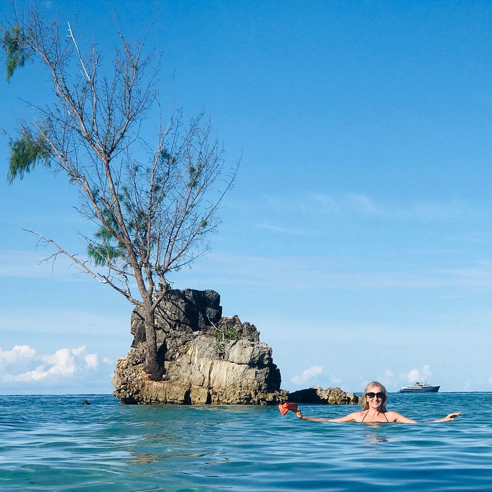 Klara Glowczewska Person enjoying a swim in clear water near a rocky islet with a single tree under a bright blue sky.