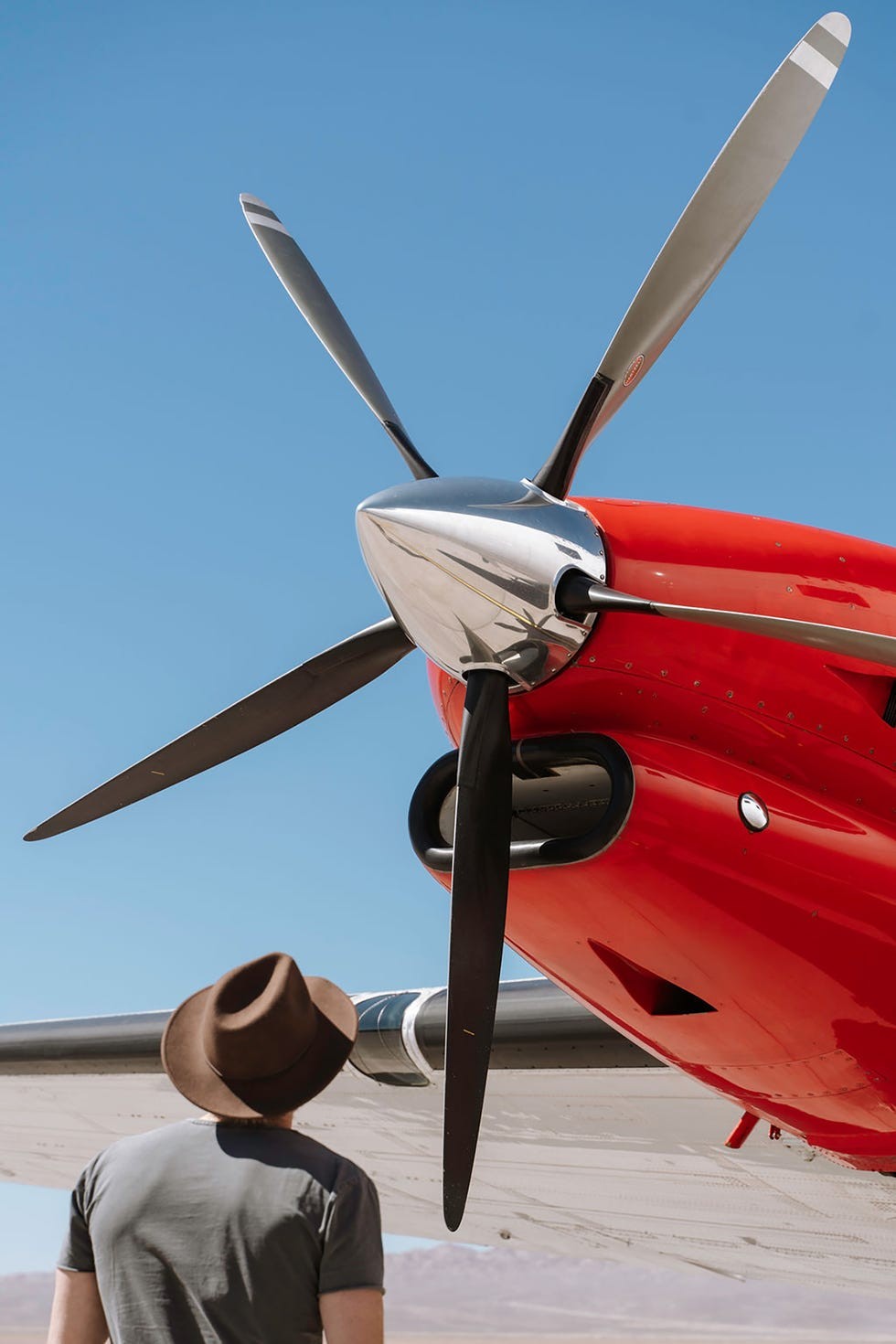 Echo Charlie expedition plane Person wearing a hat gazing up at the propeller of a bright red airplane against a clear blue sky.