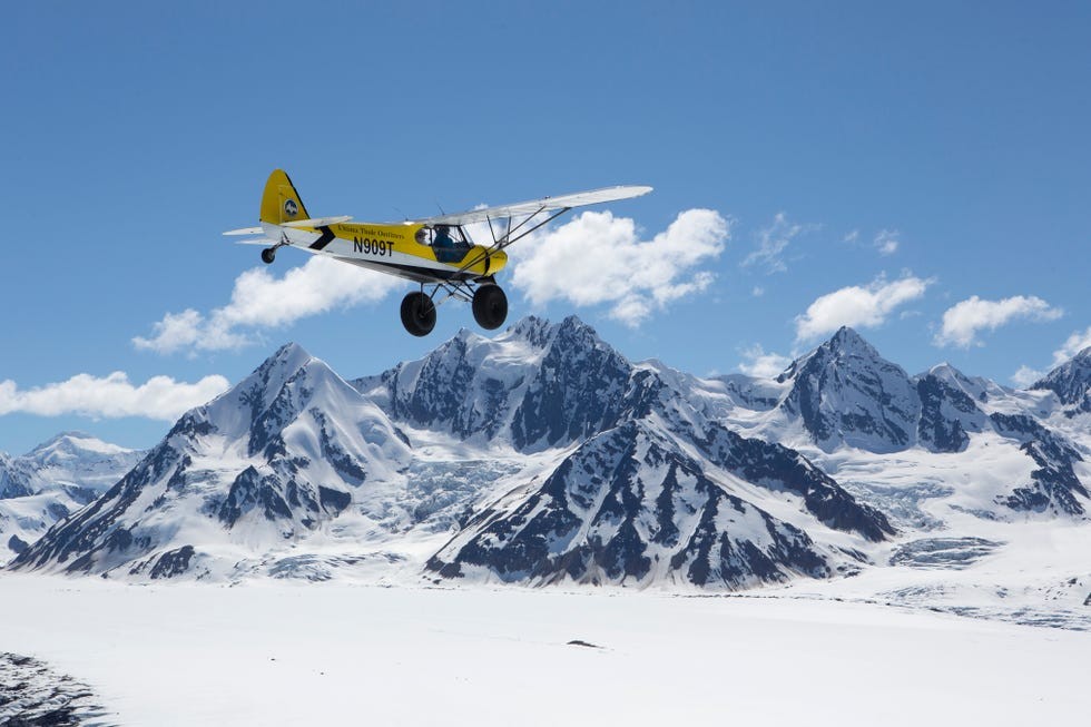 Transport at Ultima Thule Lodge in Alaska Yellow airplane flying over snowy mountains under a blue sky with scattered clouds.