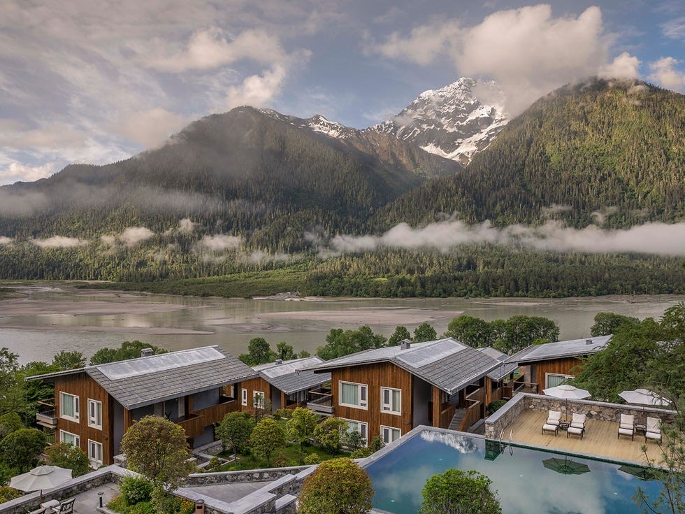 Songtsam hotels' Bome Lodge Tibet Scenic view of modern accommodations beside a river, with mountains and mist in the background.