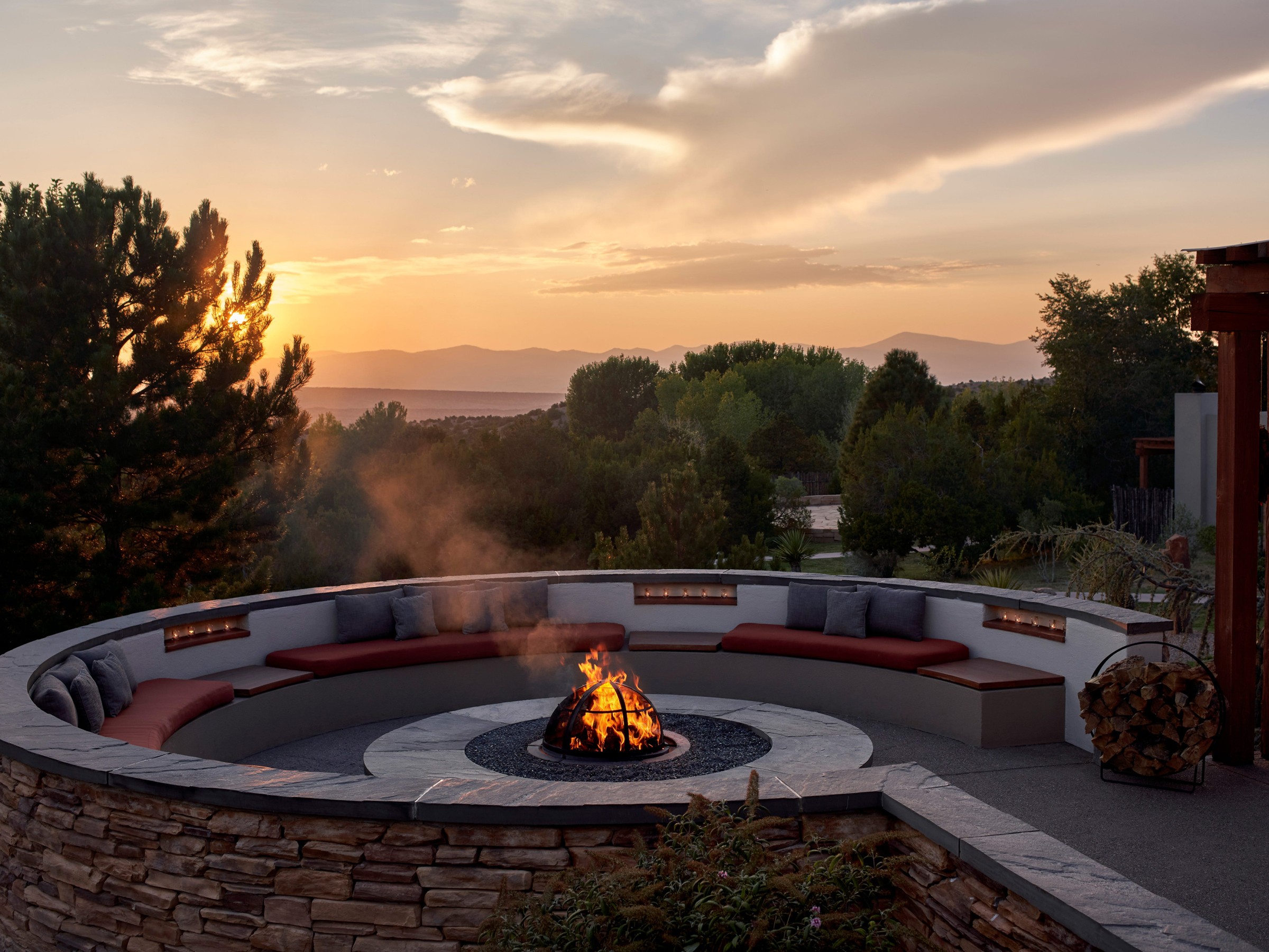 Circular seating area around a fire pit, surrounded by trees and mountains at sunset.