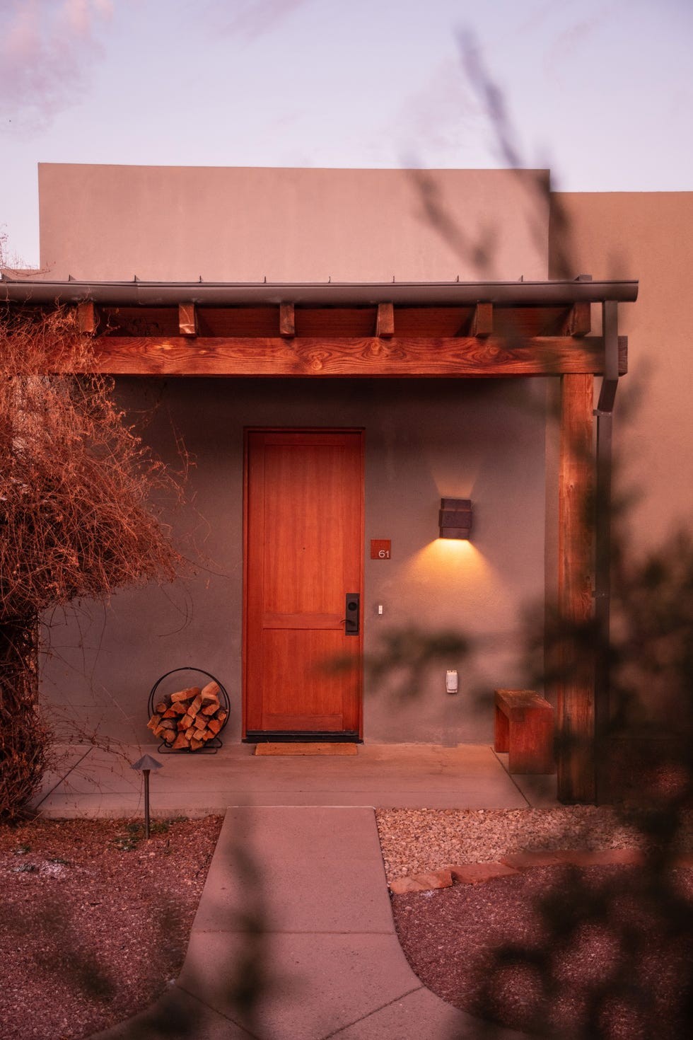 Front entrance of a modern house with a wooden door, outdoor light, and a stacked wood holder on a gravel path.