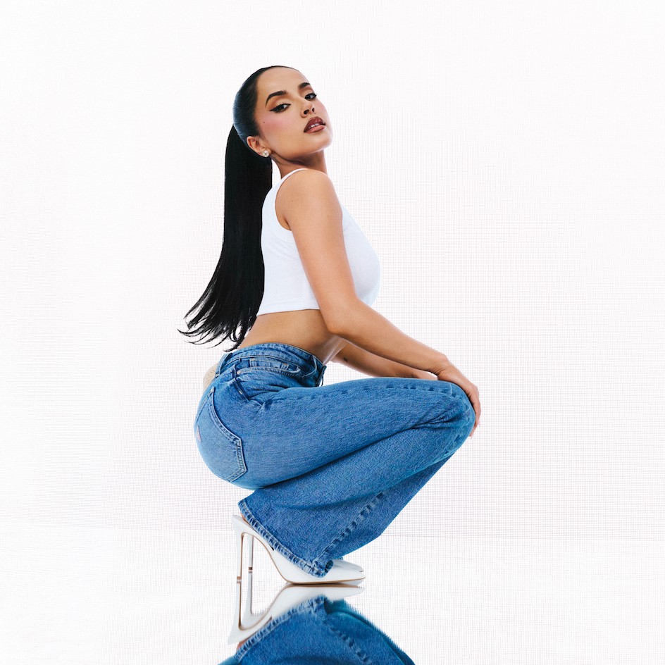 Woman in a white crop top and blue jeans crouching with long hair, reflecting in a surface beneath her.