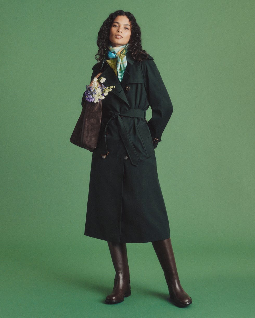A woman in a dark green coat and scarf, holding a bag with flowers, poses against a green backdrop.