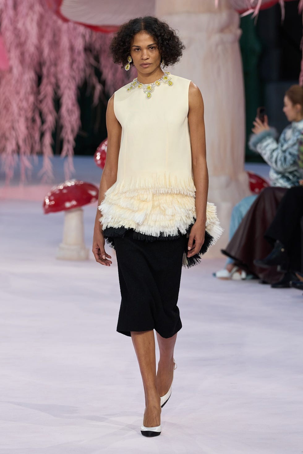 Model walking on a runway in a stylish layered outfit featuring a fringed top and black skirt, set in a colorful backdrop.