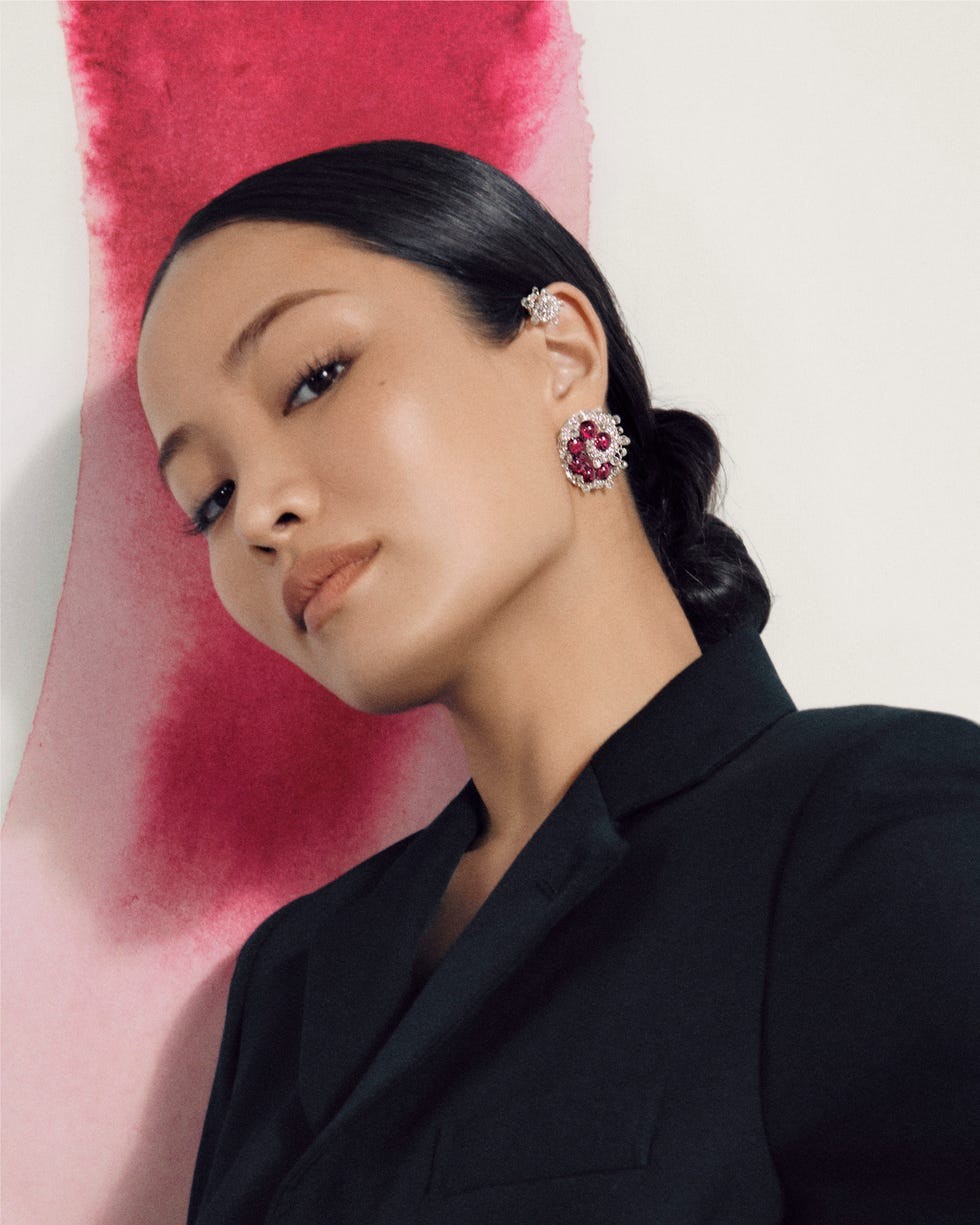 A woman wearing elegant earrings and a black blazer, posed against a backdrop of pink watercolor.
