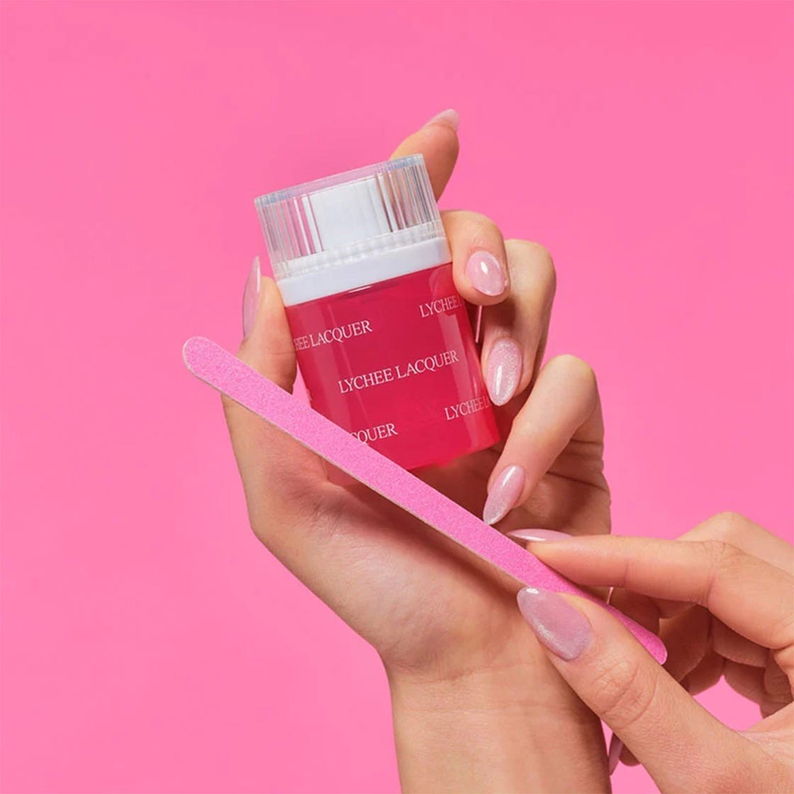 Bright pink nail polish bottle held with manicured hands and a pink nail file against a pink background.