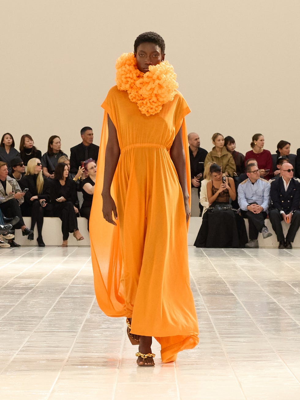 Model walking the runway in a flowing orange dress and a dramatic orange scarf, surrounded by an audience in stylish attire.