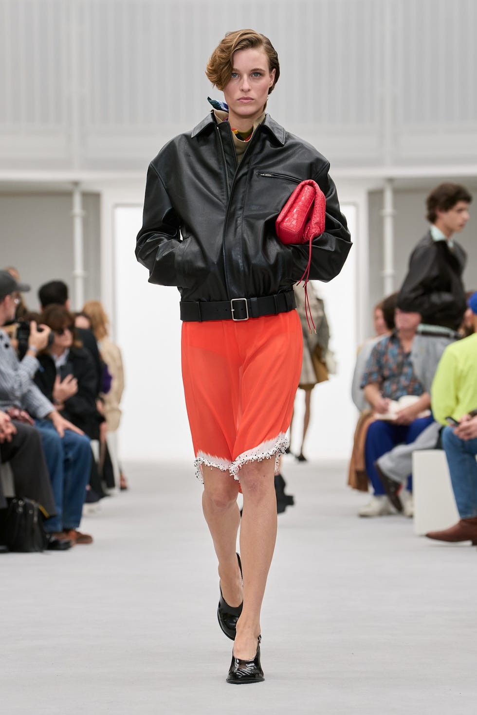 A model walks the runway in a black leather jacket, orange skirt, and black shoes during a fashion show.