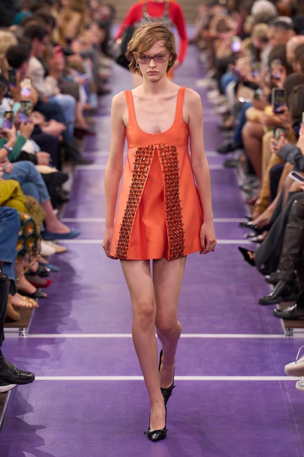 A model walks the runway wearing an orange dress with embellishments during a fashion show.