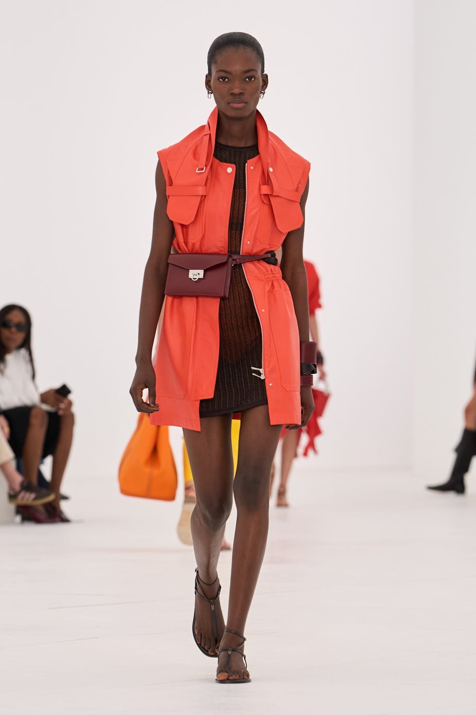Model walking on runway wearing an orange outfit and carrying a small bag, with audience visible in the background.