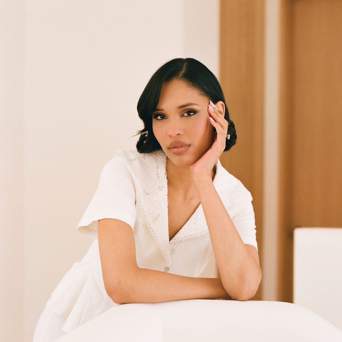 Young woman in a white outfit resting her chin on her hand, set against a light-filled, minimalist background.