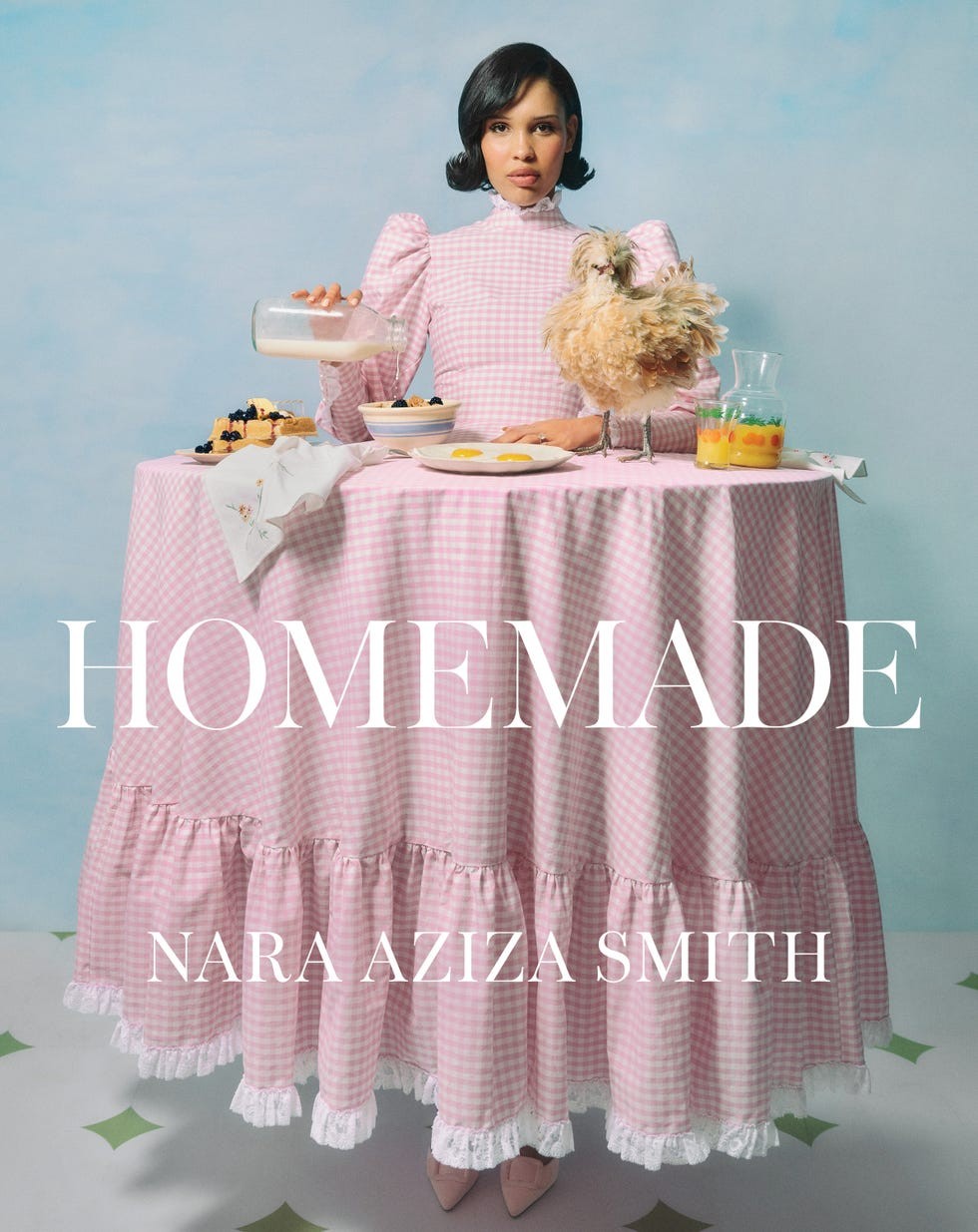 A woman in a pink checkered dress pours milk while surrounded by a table setting featuring breakfast foods and a chicken.