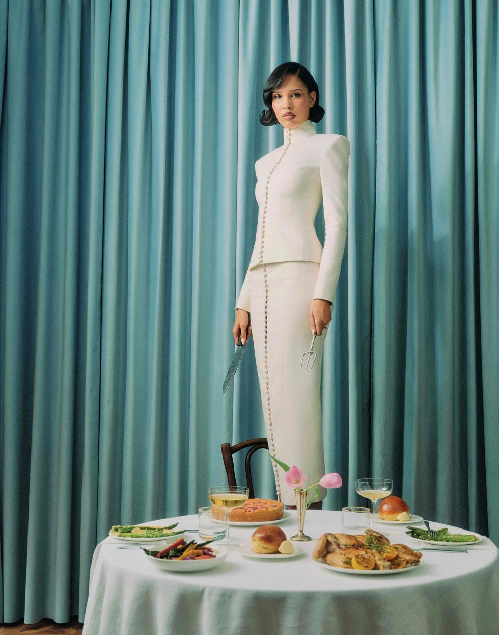 A poised model in a stylish suit stands by a elegantly laid table with diverse dishes and drinks against a pale blue backdrop.
