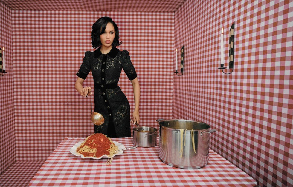 A woman in a black lace dress serves spaghetti from a pot in a checkered room.