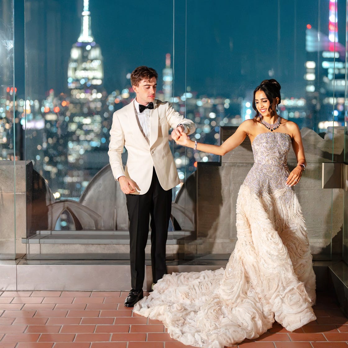 A couple elegantly dressed, holding hands with a city skyline and the Empire State Building in the background at night.