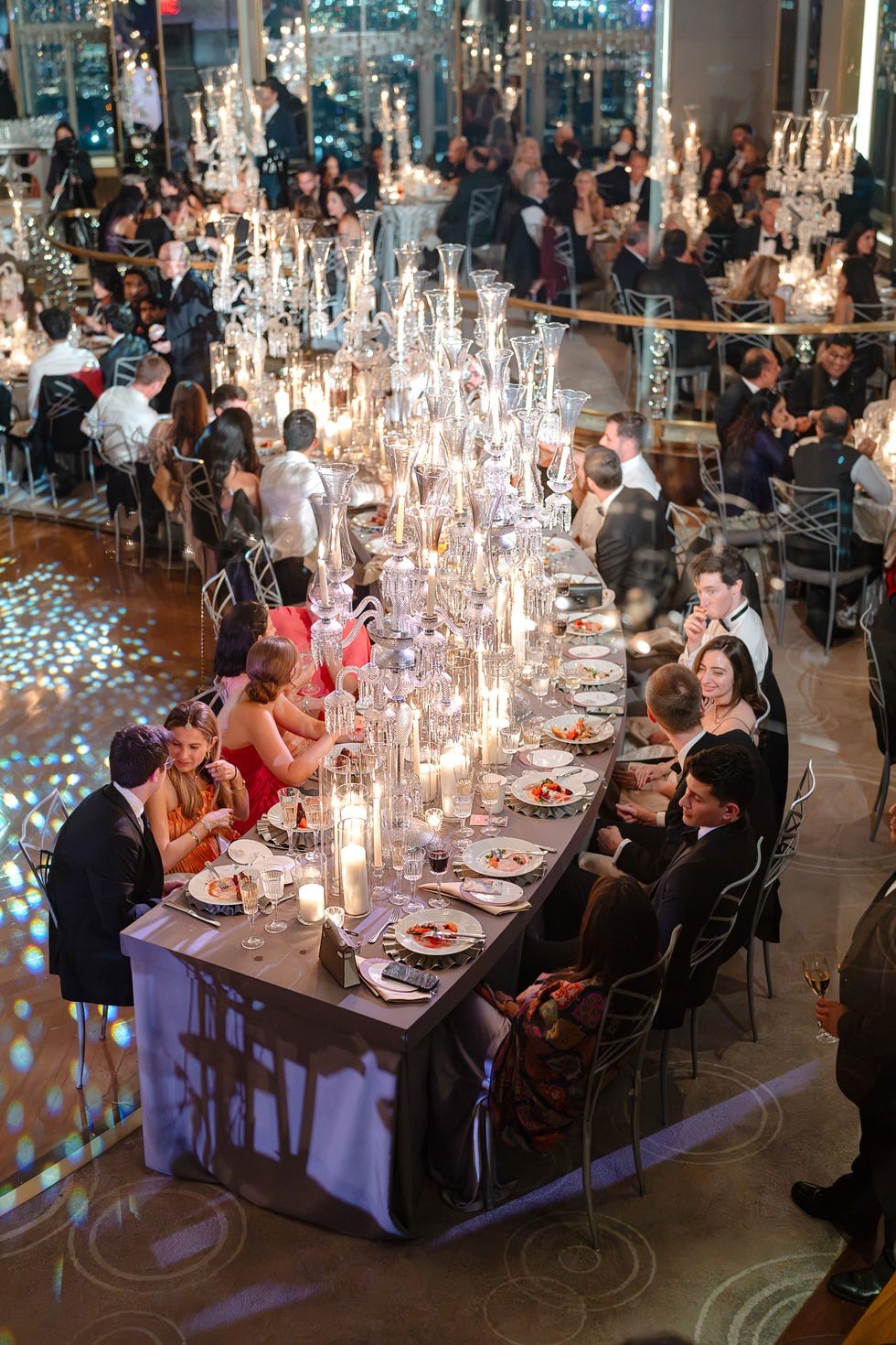 Elegant dining setup with guests enjoying a festive event, adorned with candelabras and reflective decor.