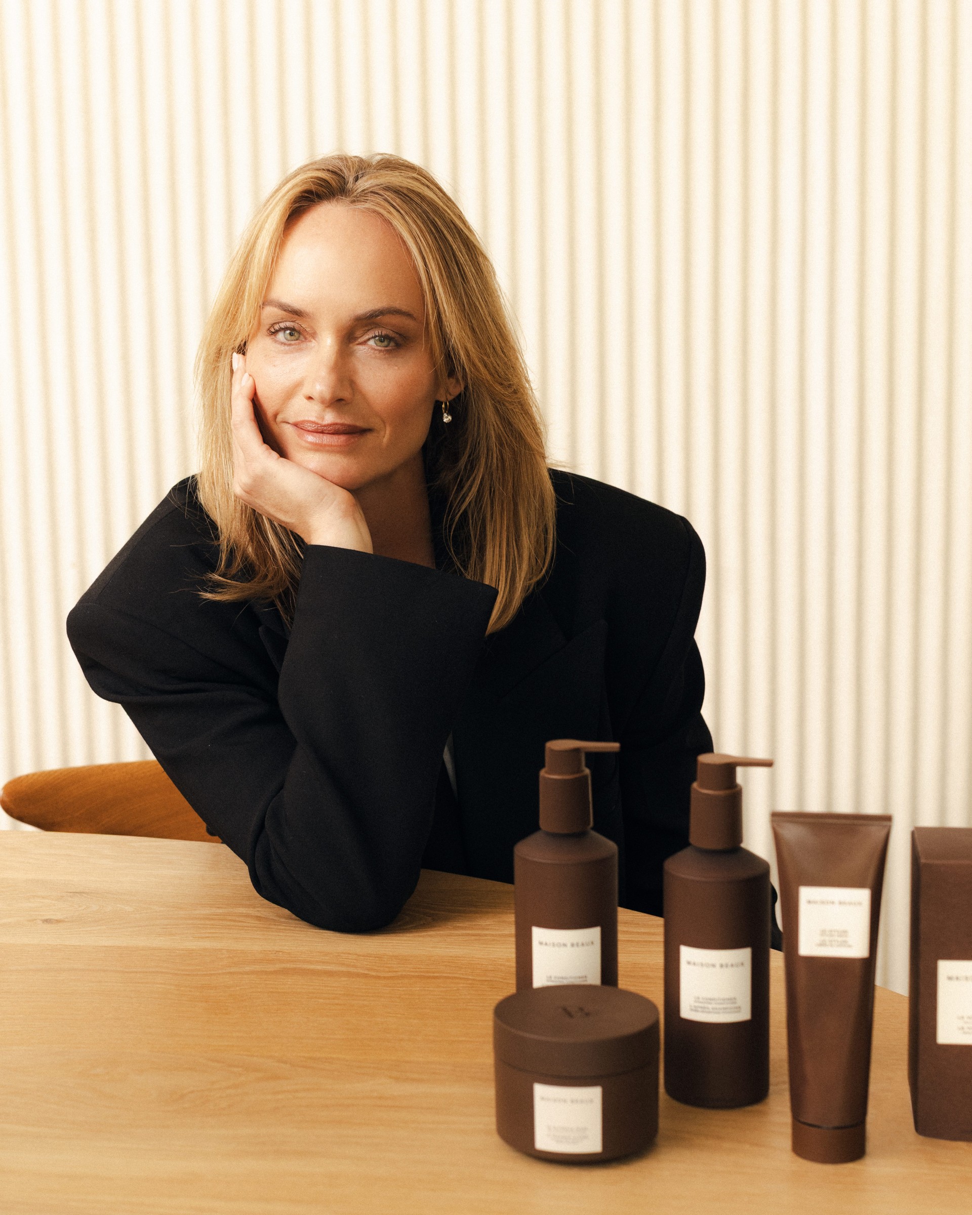 A woman in a black blazer sits at a wooden table with various skincare products displayed.