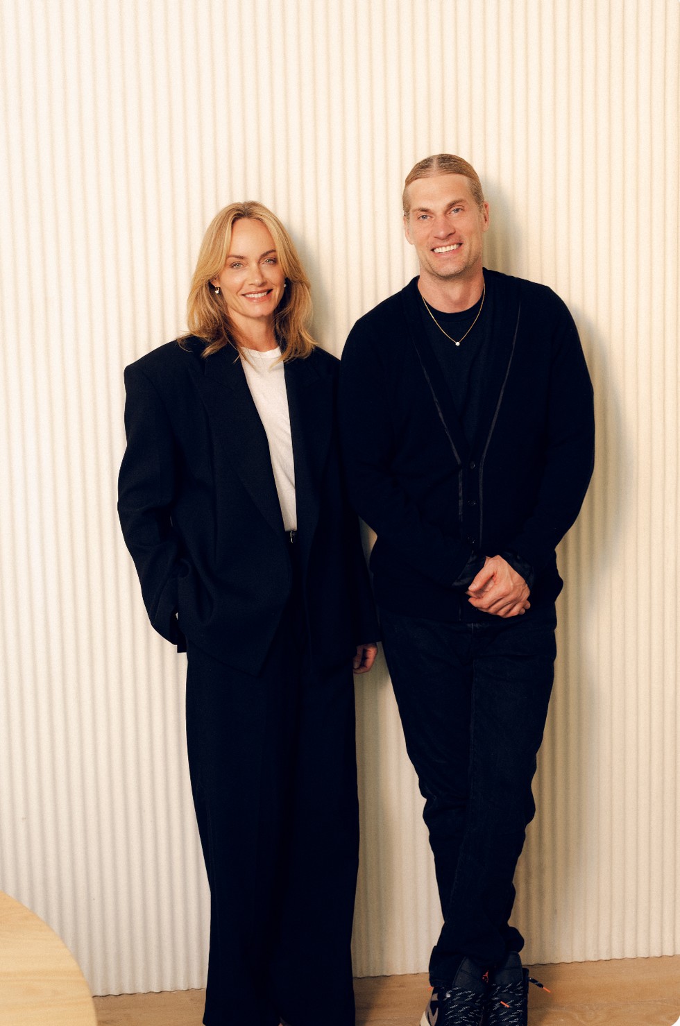 Two smiling individuals posing together, one in a black suit and the other in a black cardigan with jeans.