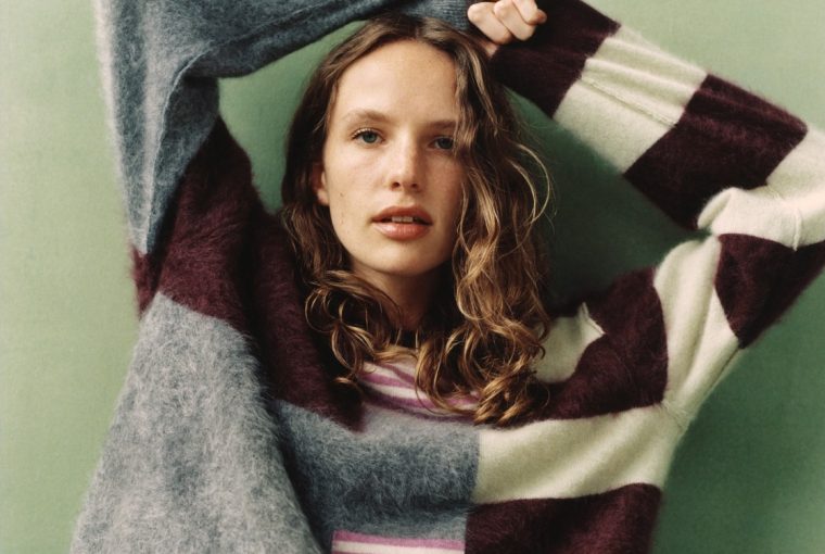 A woman with curly hair poses in a colorful, patterned sweater against a green background.