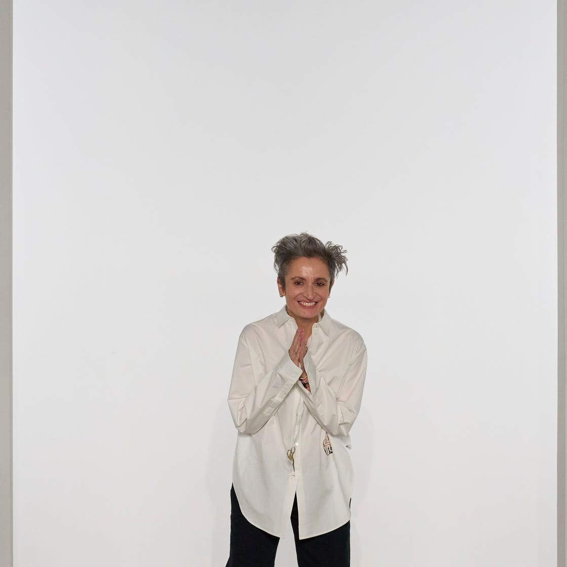 A smiling person in a white shirt with hands together in a gesture of greeting, standing in an art gallery.