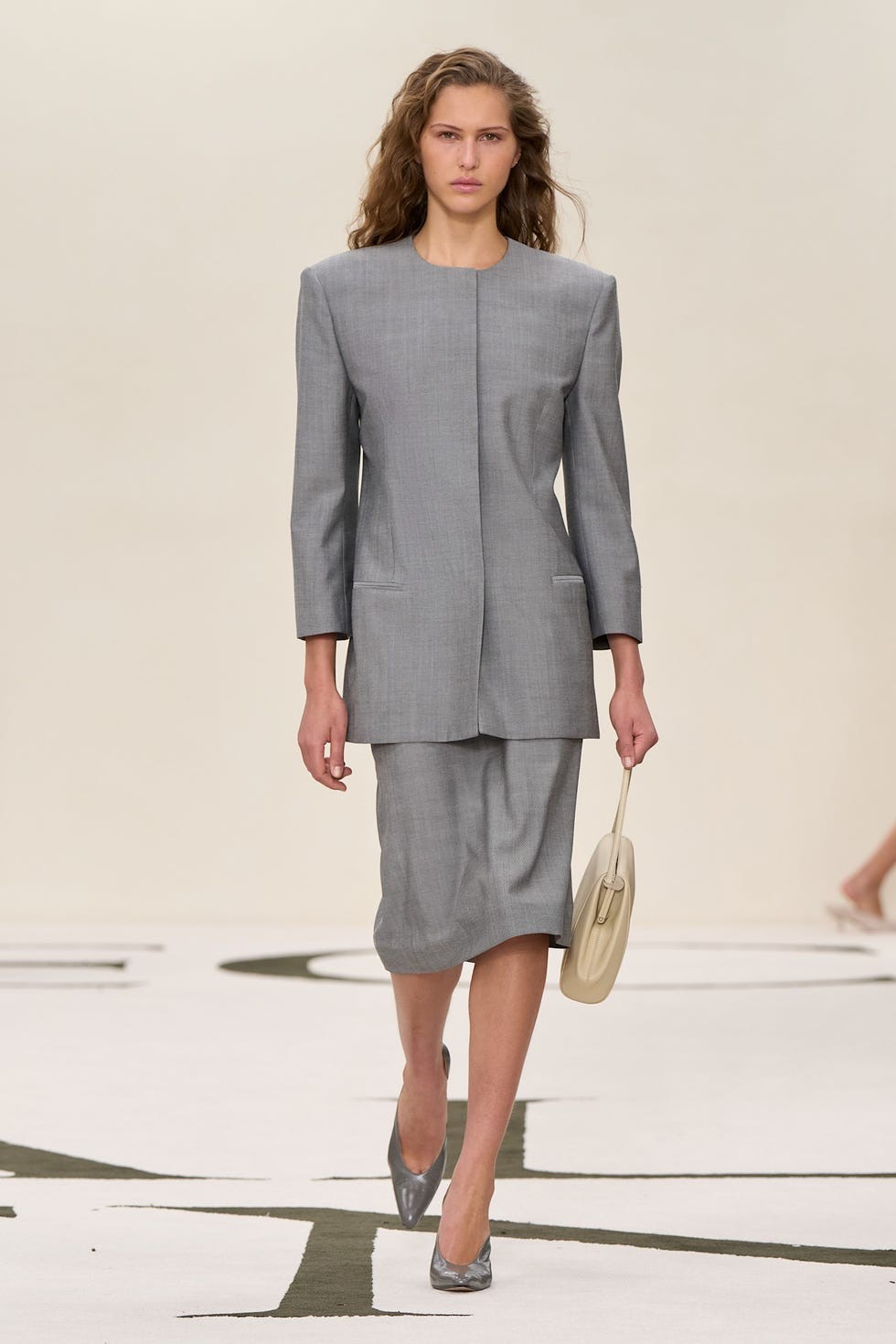 Model walking on runway in a stylish gray suit with a matching skirt and gray heels, holding a beige handbag.