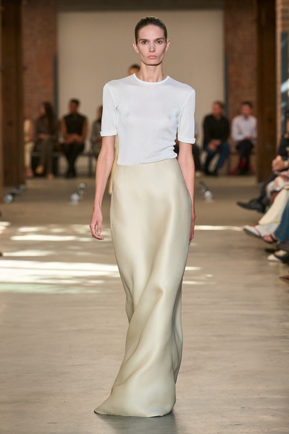 Model walking on the runway wearing a fitted white top and a flowing off-white skirt during a fashion show.