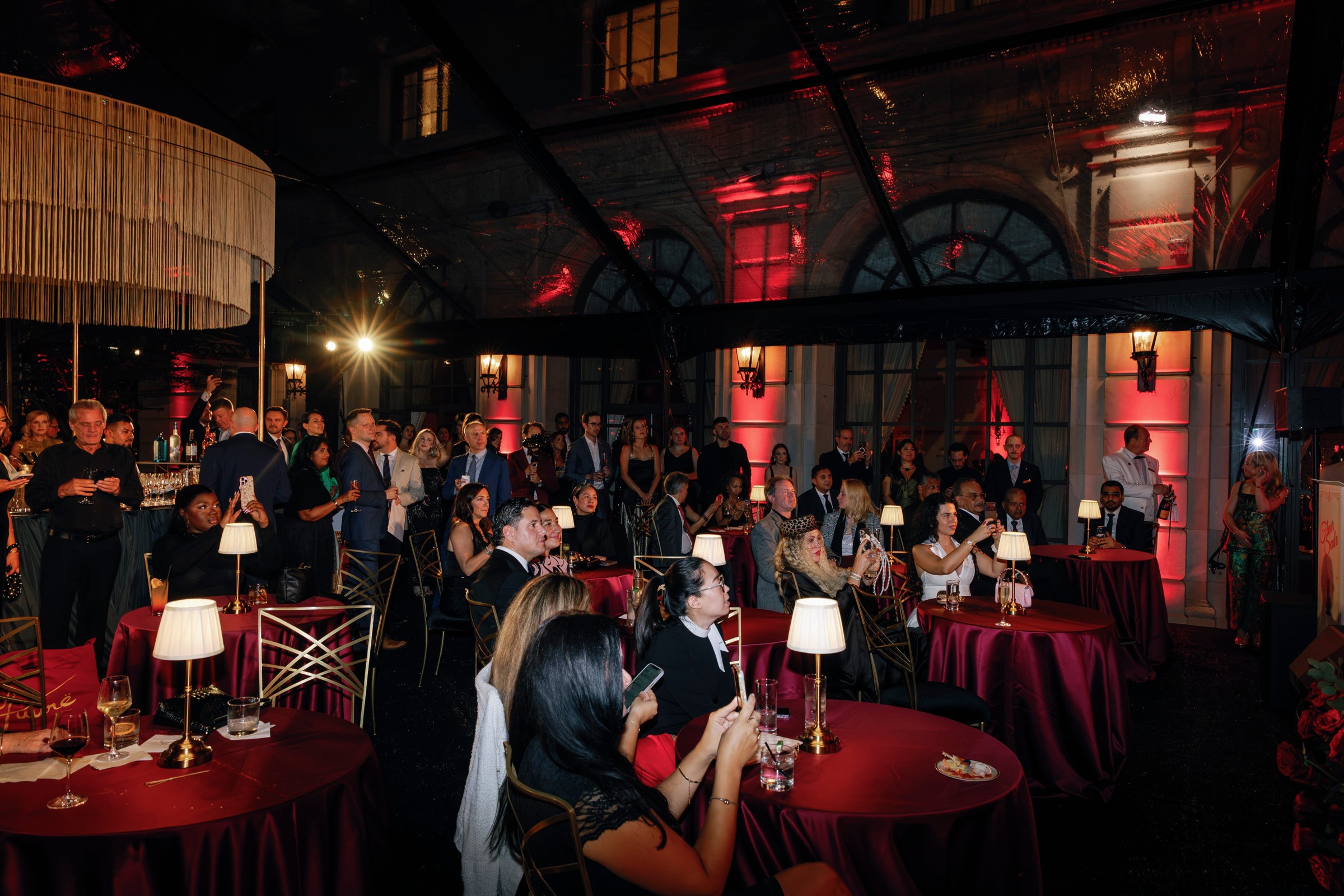 A lively event with guests socializing around tables, enjoying drinks and entertainment under soft lighting.