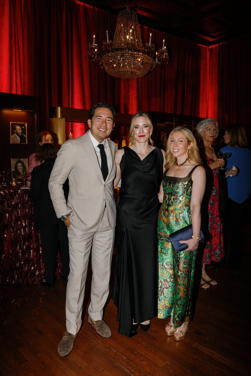 Three individuals posing together at a stylish event with a chandelier and red drapery in the background.
