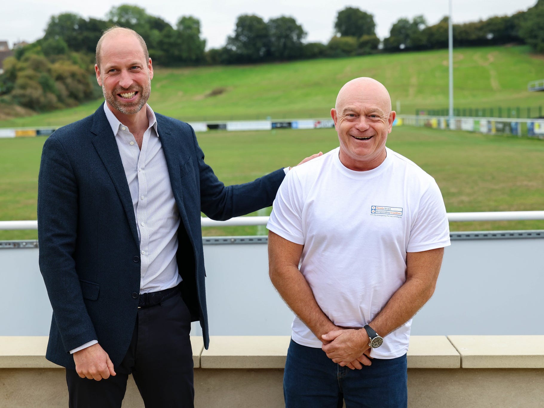 Two men smiling together at a sports field with greenery and an empty stand in the background.