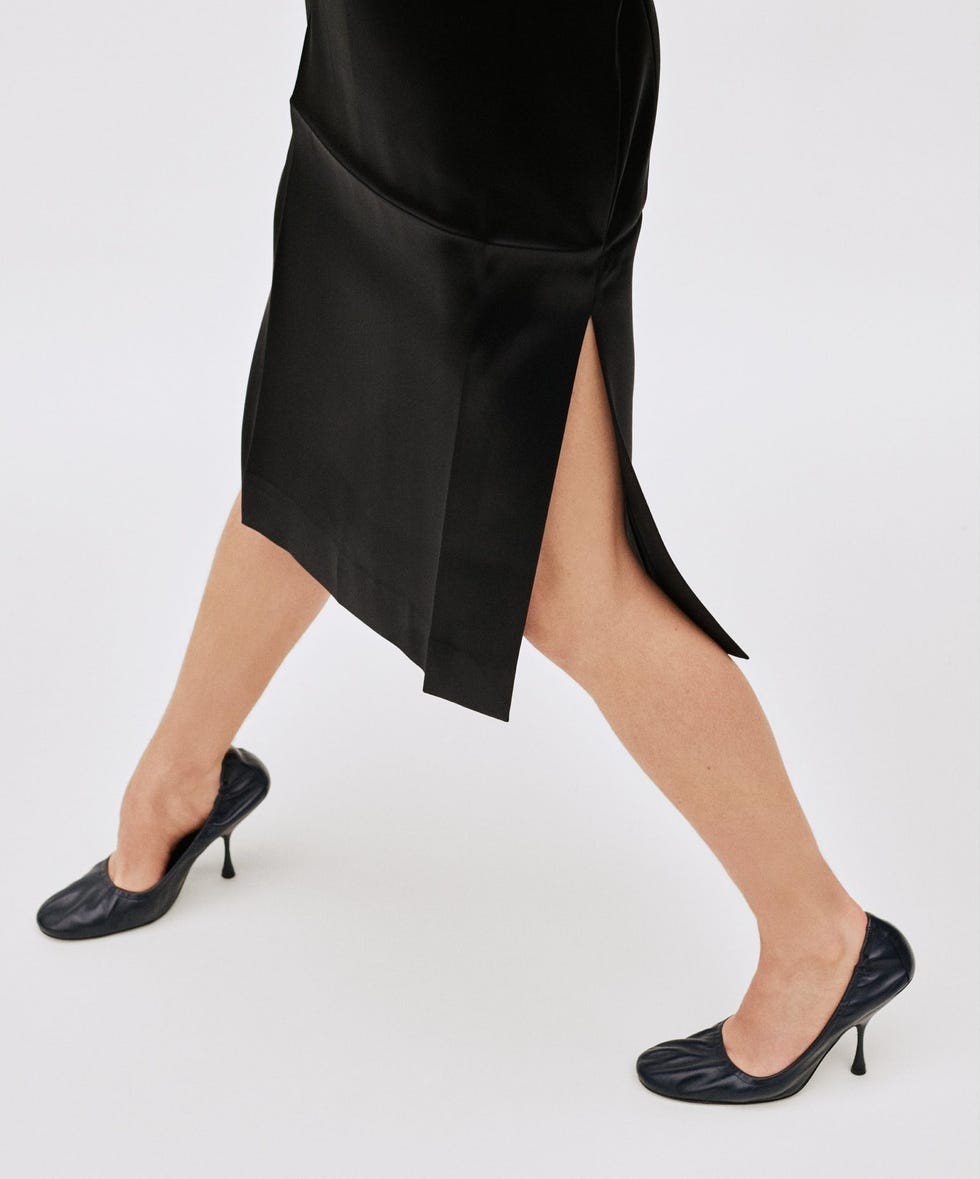 Leg in a black satin dress with a side slit, paired with black high-heeled shoes on a light background.