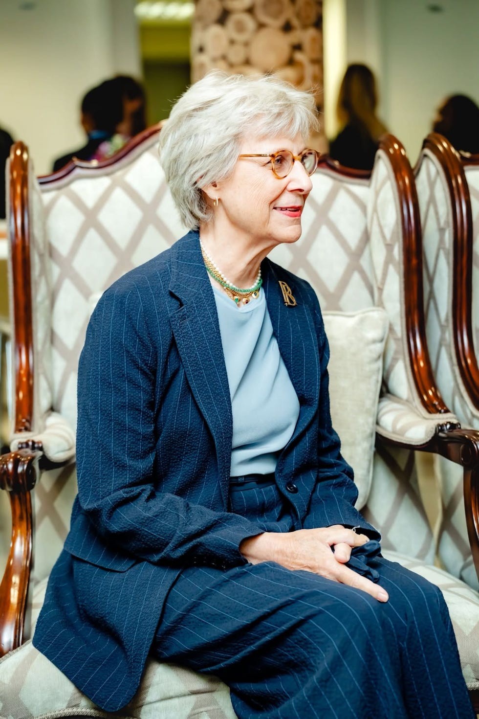 Older woman with glasses wearing a blue outfit seated in an ornate chair, smiling in a well-lit indoor setting.