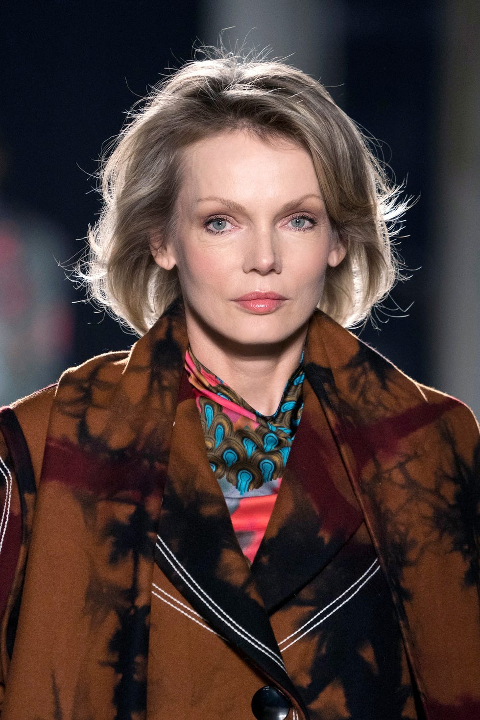 Person with short blonde hair wearing a patterned scarf and brown coat