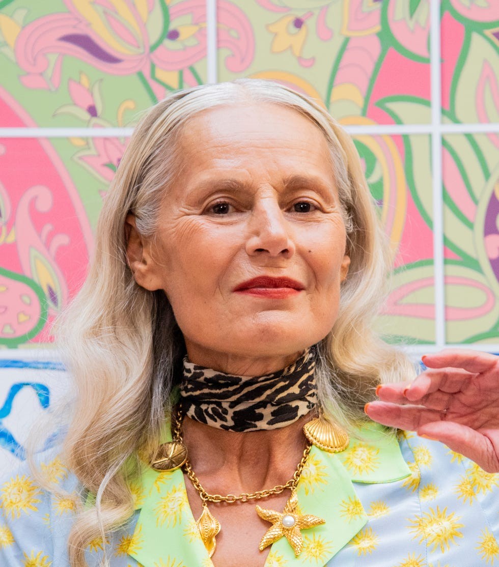 Elderly woman with long hair and vibrant patterned outfit in front of colorful background.