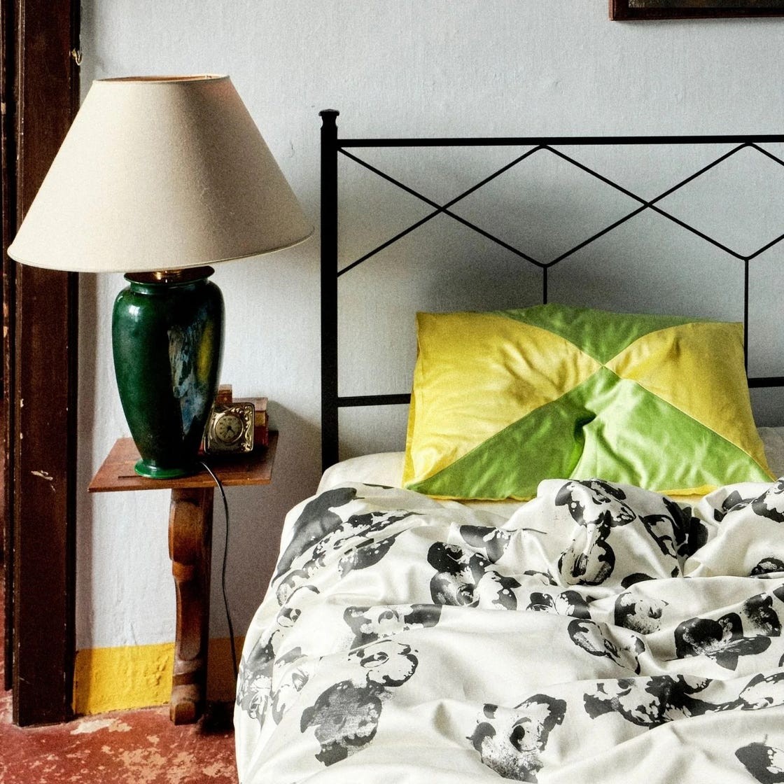 Cozy bedroom scene with a decorative lamp, colorful pillows, and a patterned blanket on an iron bedframe.