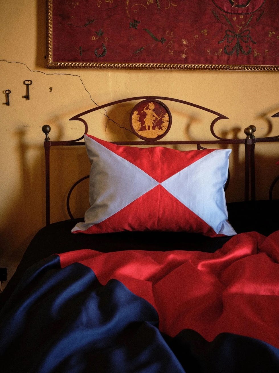 A bed with a decorative pillow featuring red and blue geometric patterns, alongside a richly colored blanket.