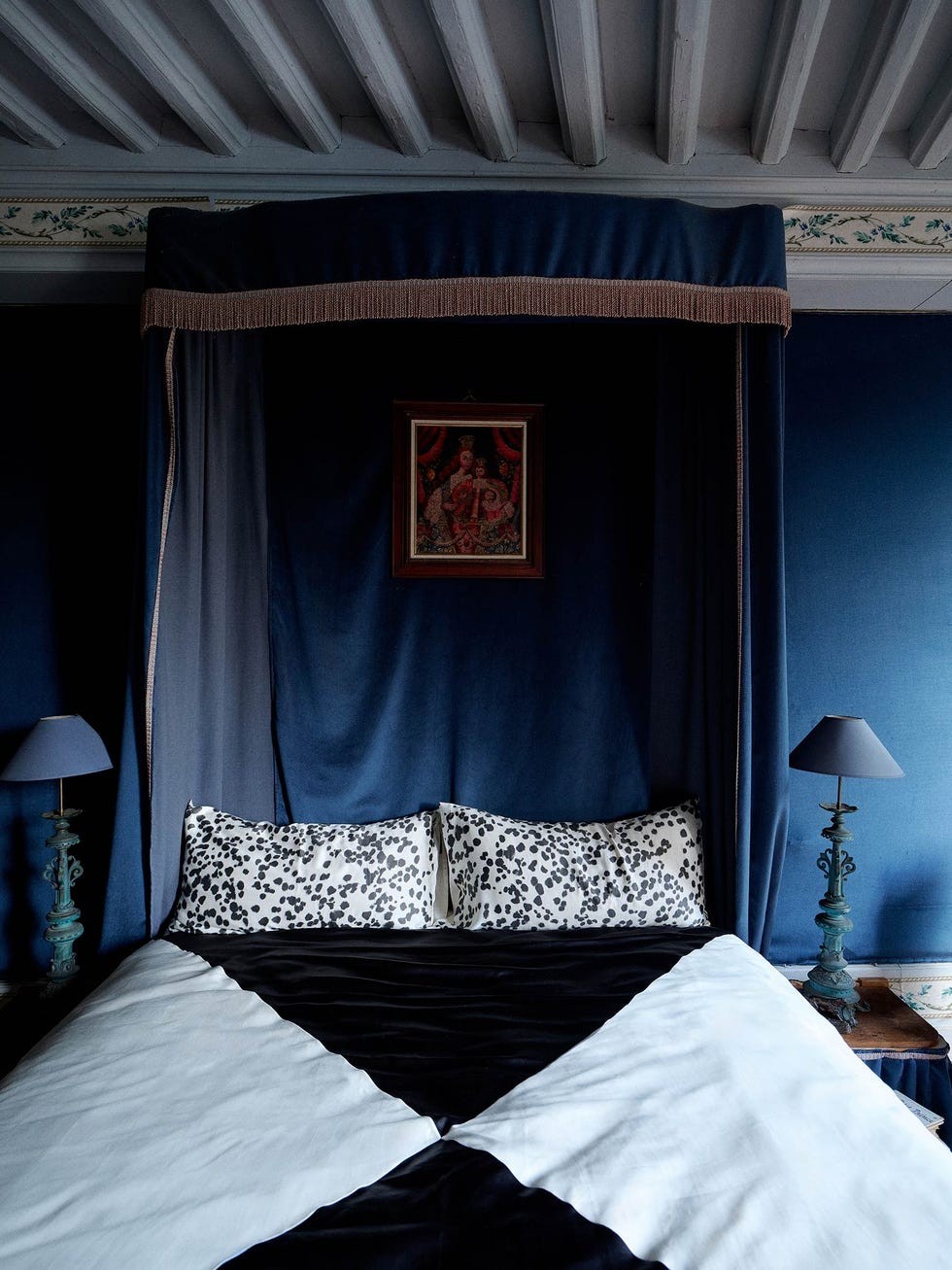 A stylish bedroom featuring a dark canopy, patterned pillows, and a vintage lamp against blue walls.