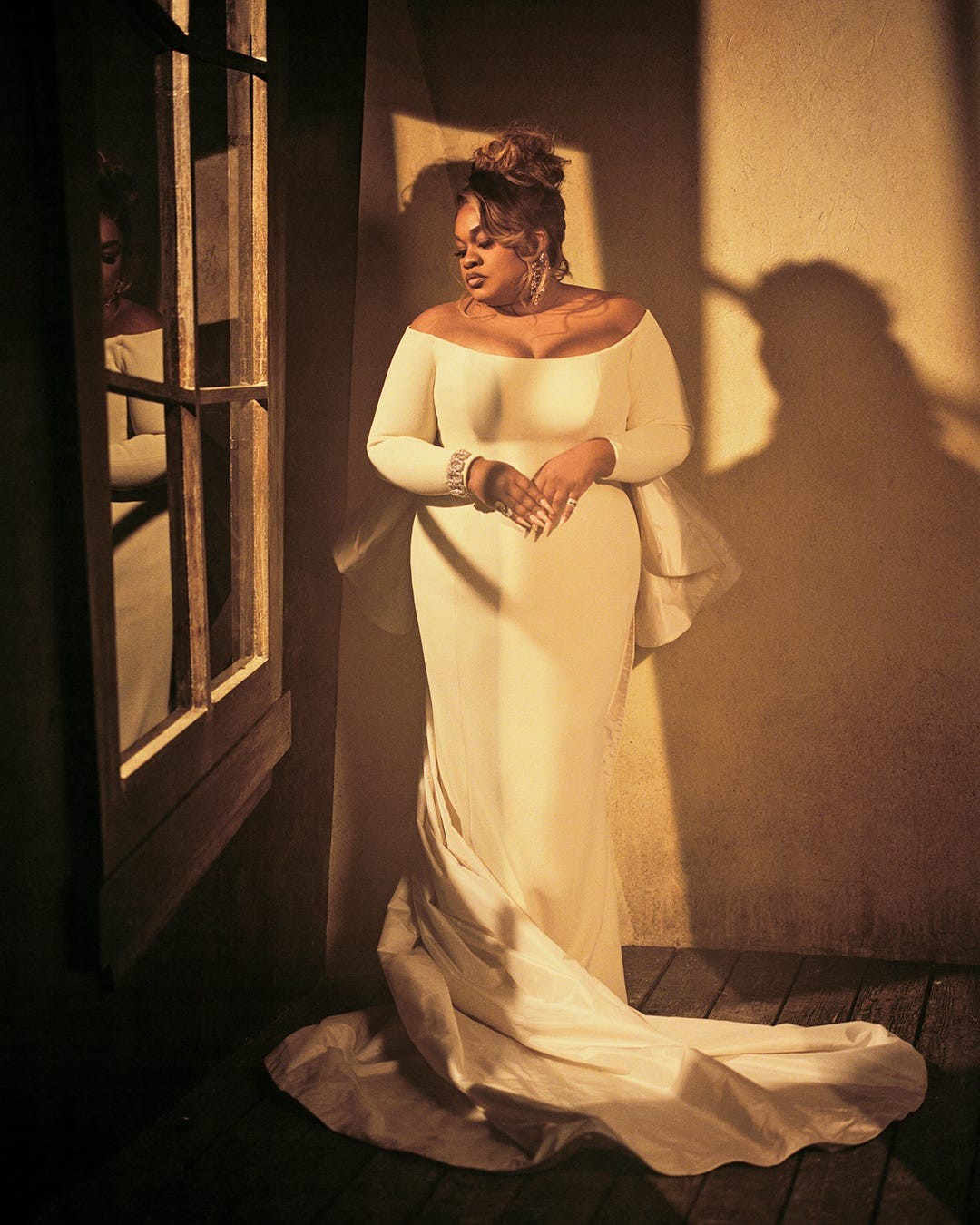 A woman in an elegant off-shoulder gown stands gracefully by a window, casting a soft shadow in warm light.