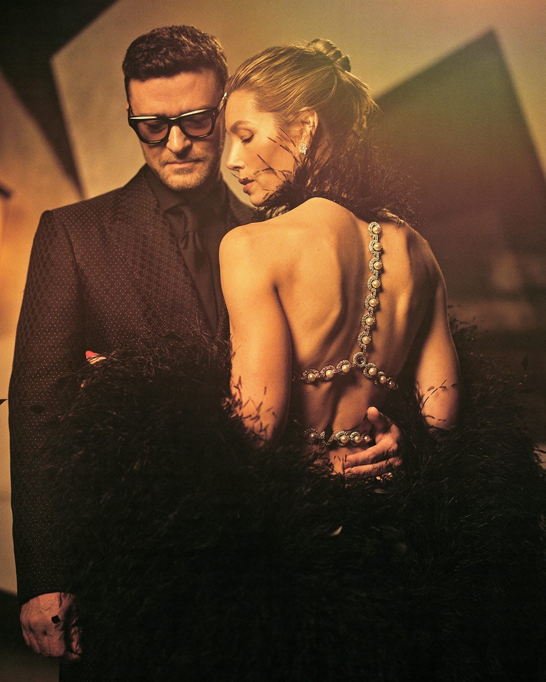 A stylish couple embraces, showcasing elegant fashion and a dramatic feather boa in a warm, artistic setting.