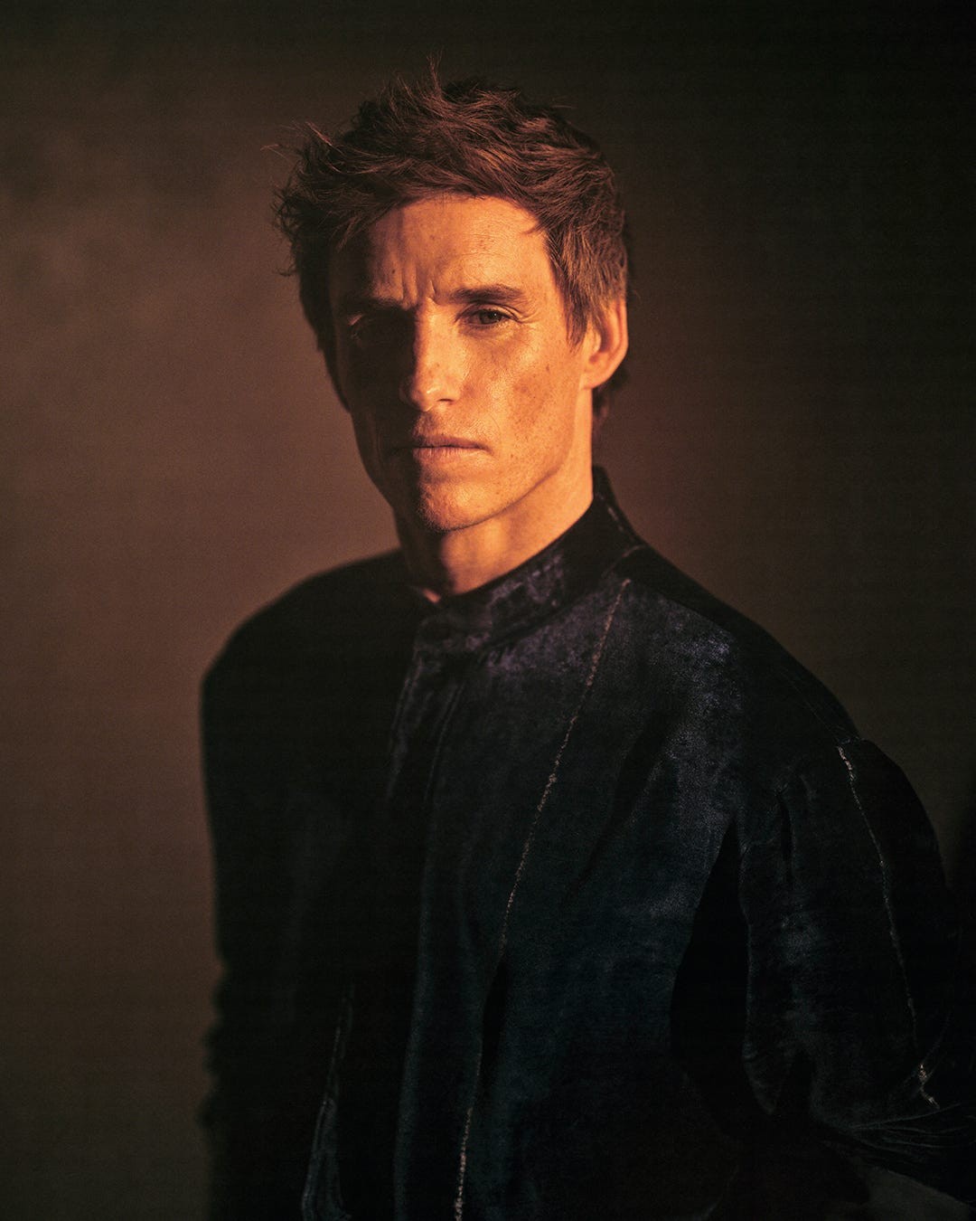 A man in a dark velvet shirt gazes intently, illuminated by soft, warm lighting.
